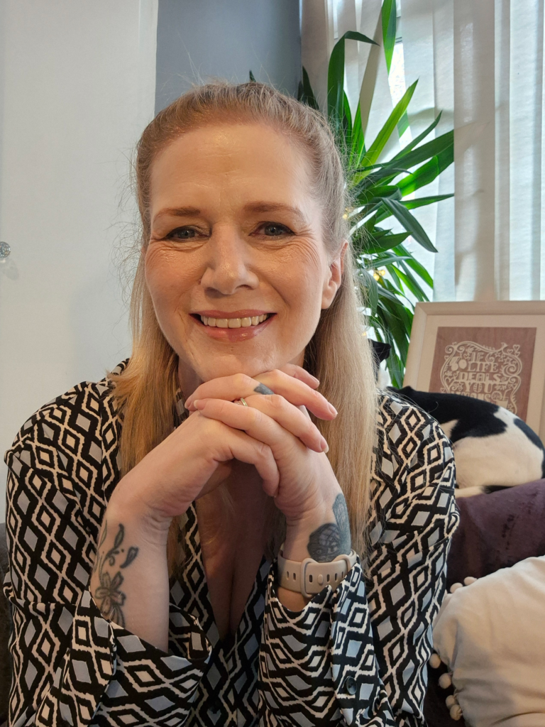 Kate Stapleton, a blonde woman, is smiling at the camera with fingers interlaced and wearing a patterned shirt. She is in front of a yukka plant.